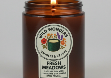Candle in an amber jar with a label reading 'Wild Wonders Candles & Crafts Fresh Meadows' on a light gray background.