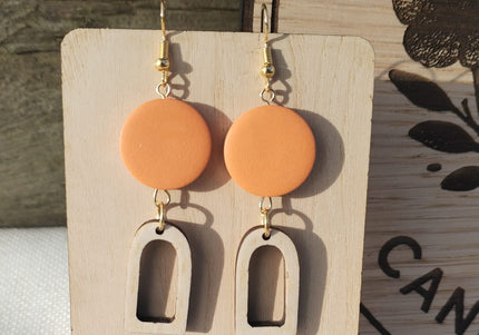 Pair of orange earrings with wooden arches on a wooden stand with purple flowers.