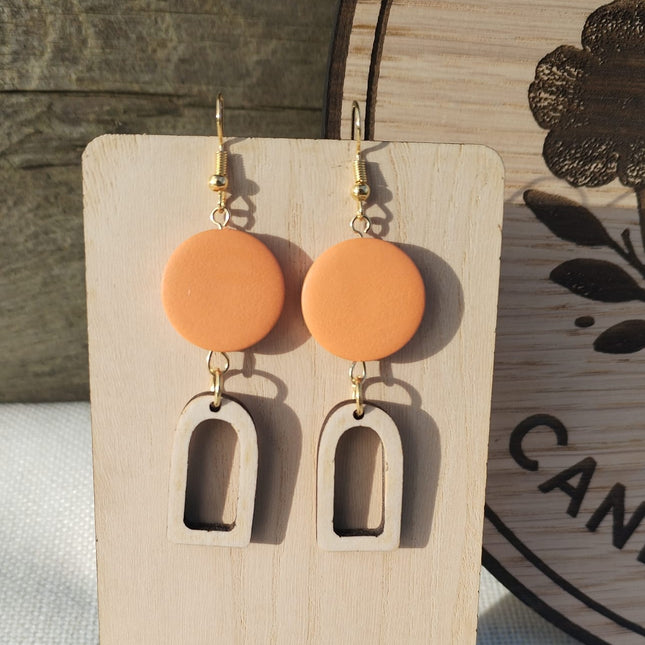 Pair of orange earrings with wooden arches on a wooden stand with purple flowers.