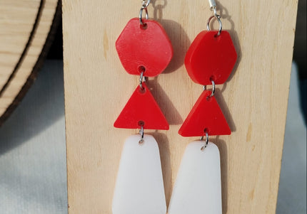 Red and white geometric earrings on a wooden stand with flowers.