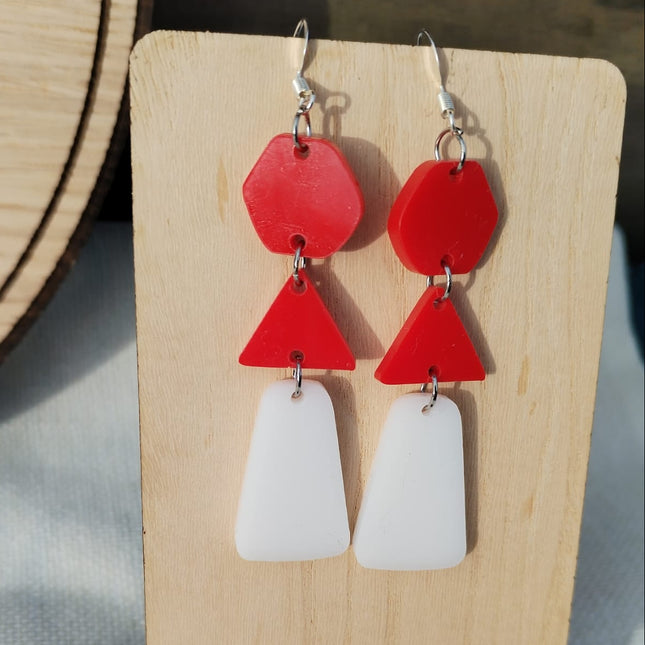Red and white geometric earrings on a wooden stand with flowers.
