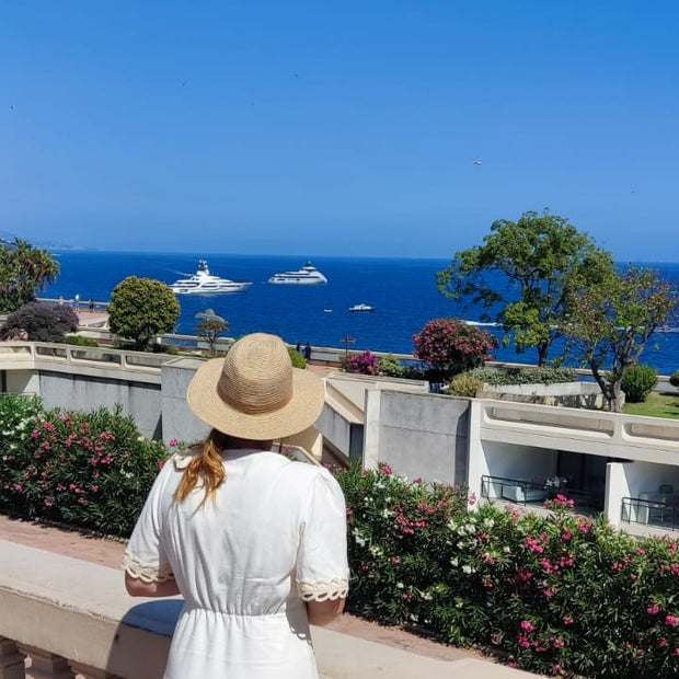 Woman in a white dress and hat overlooking a scenic coastal view with boats on the water.